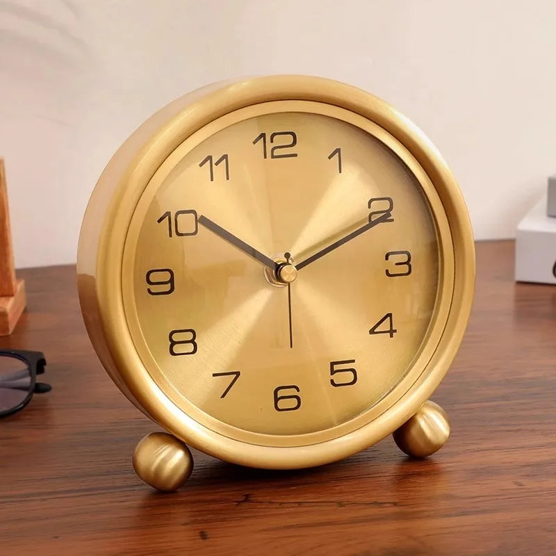 Brass Table Clock - Helric: A round, gold-colored analog clock with black numerals and hands, resting on ball feet, exuding timeless elegance amidst eyeglasses and books on a wooden surface.