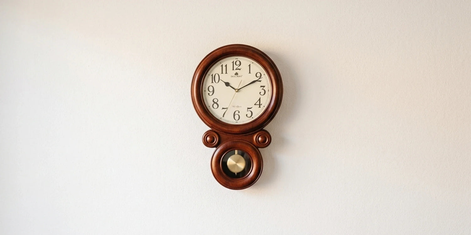 A light-beige sofa sits by a window with matching curtains. Above a wooden sideboard, a house-shaped pendulum clock is mounted, complemented by a ceramic lamp and vase with dried grasses.