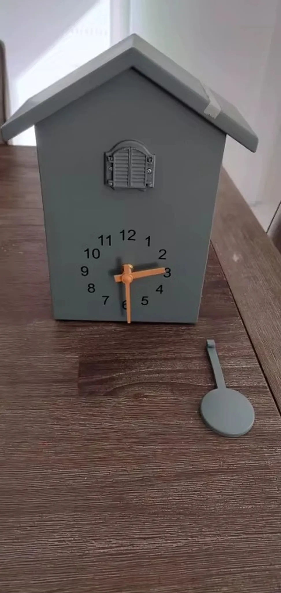 Wooden Cuckoo Clock - Brivon: A matte gray, house-shaped tabletop clock with a printed face, black numbers, and bright orange hands, featuring an unhooked gray pendulum and shuttered window above the dial.