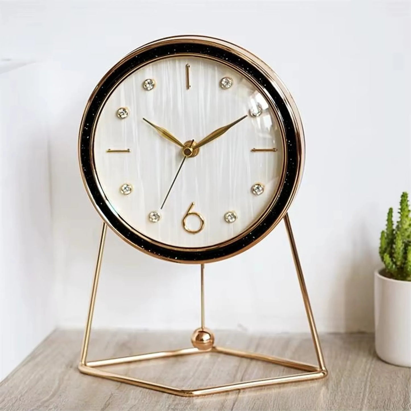The Dralen Pendulum Table Clock features a white face with gold accents, a minimalist gold-wire stand, and a swinging pendulum, complemented by a potted succulent for a modern, elegant display.