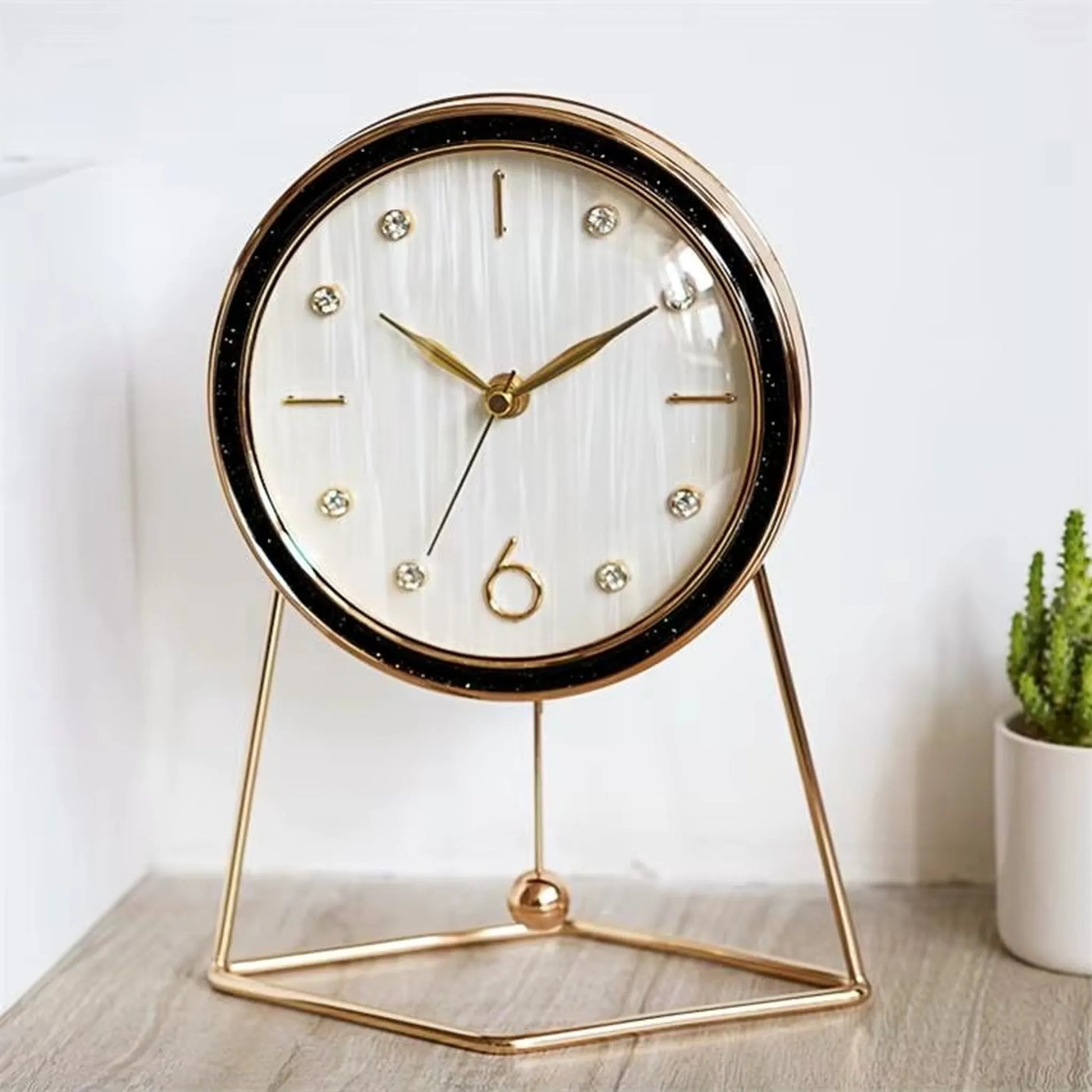 The Dralen Pendulum Table Clock features a white face with gold accents, a minimalist gold-wire stand, and a swinging pendulum, complemented by a potted succulent for a modern, elegant display.
