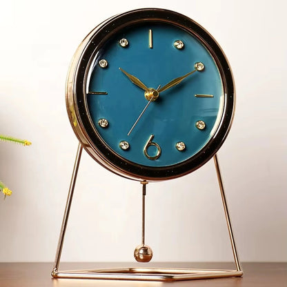 Pendulum Table Clock - Dralen features a teal-blue dial with gold hands, crystal markers, and a stylized 6, framed in gold with a triangular stand and hanging pendulum for elegant decor.