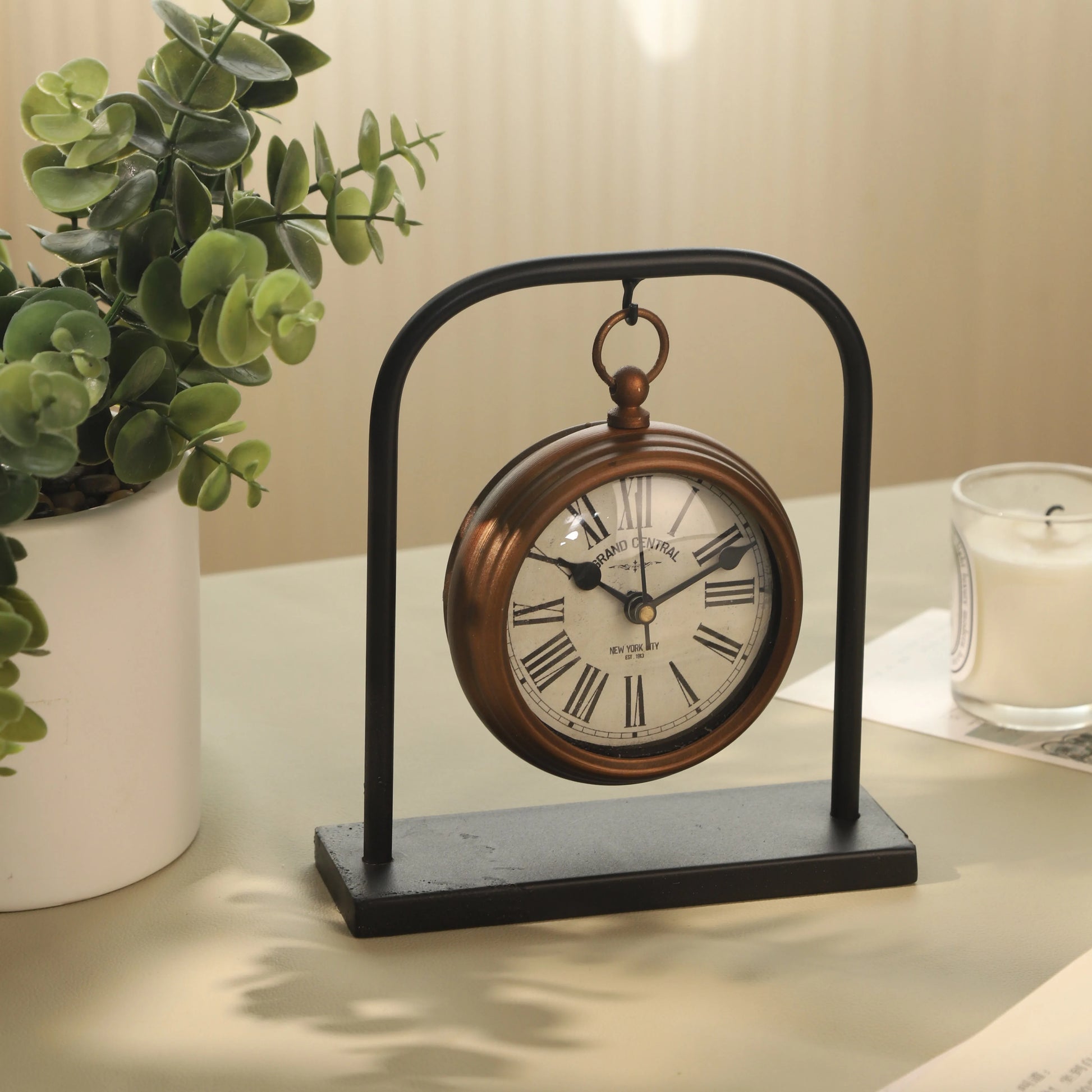 Retro Table Clock - Kelros: A vintage-style tabletop clock with a bronze-toned case and Roman numerals, elegantly suspended on a black metal arch, perfect for enhancing a desk or bedroom décor.