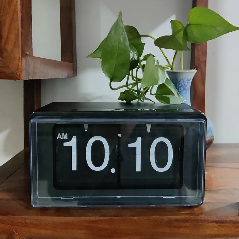 Vintage flip clock - TimeFlip displayed in a clear plastic case on a wooden shelf, reading “AM 10:10.” A small potted plant in a blue-and-white ceramic vase complements the retro aesthetic.