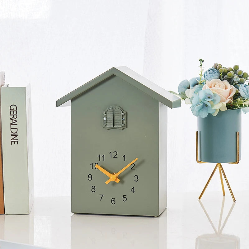 Wooden Cuckoo Clock - Brivon: A charming house-shaped tabletop clock with bright yellow hands, featuring a faux window detail. Flanked by a book stack and pastel flowers, adding modern charm.