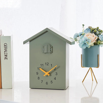 Wooden Cuckoo Clock - Brivon: A charming house-shaped tabletop clock with bright yellow hands, featuring a faux window detail. Flanked by a book stack and pastel flowers, adding modern charm.