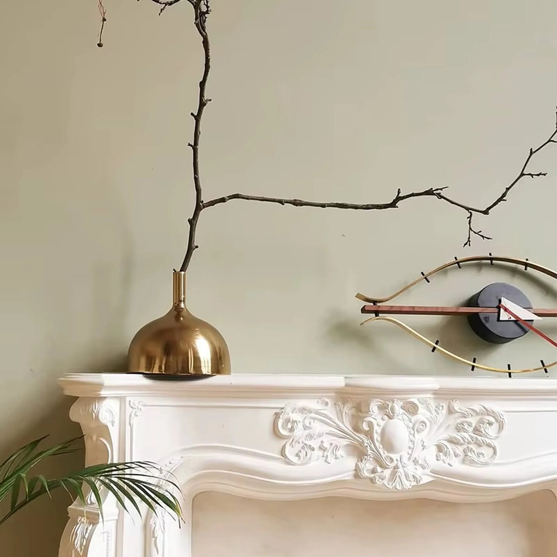 30-inch decorative wall clock - Ashen: A sculptural eye-shaped clock with gold metal rods and a straight copper-tone hand, set against a muted-green wall above a white mantel.