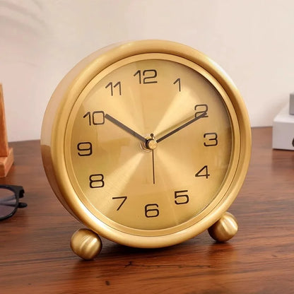 Brass Table Clock - Helric: A round, gold-colored analog clock with black numerals and hands, resting on ball feet, exuding timeless elegance amidst eyeglasses and books on a wooden surface.