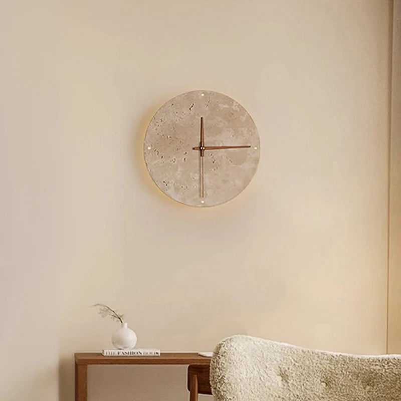 Modern wall clock for living room - Torwyn: A minimalist 9-inch travertine disc with slender metal hands, featuring natural pores and a warm LED halo, is elegantly displayed on a light-beige wall.