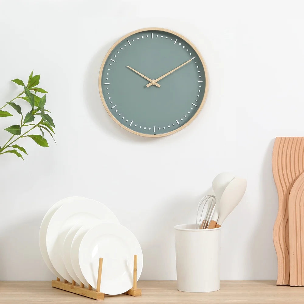 Small modern wall clock - Merrin with a muted teal face and light wood frame, mounted above a minimalist kitchen vignette featuring natural wood tones and clean white accents.