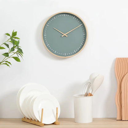 Small modern wall clock - Merrin with a muted teal face and light wood frame, mounted above a minimalist kitchen vignette featuring natural wood tones and clean white accents.