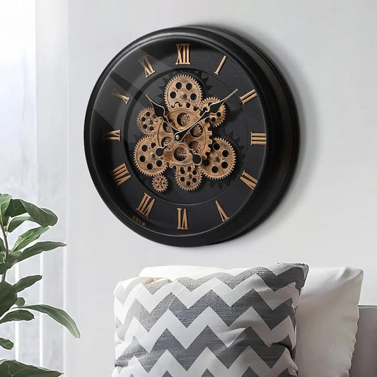 Oversized Wall Clock - Kairos, featuring a black frame with distressed finish, showcases exposed brass gears and Roman numerals. It’s mounted on a light wall, complemented by a nearby leafy houseplant.