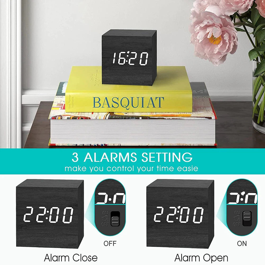 Cube Alarm Clock - Corven displayed on stacked books, features a matte-black wooden finish with white LED numerals. Three alarm settings highlighted on a teal banner, vase of flowers adjacent.