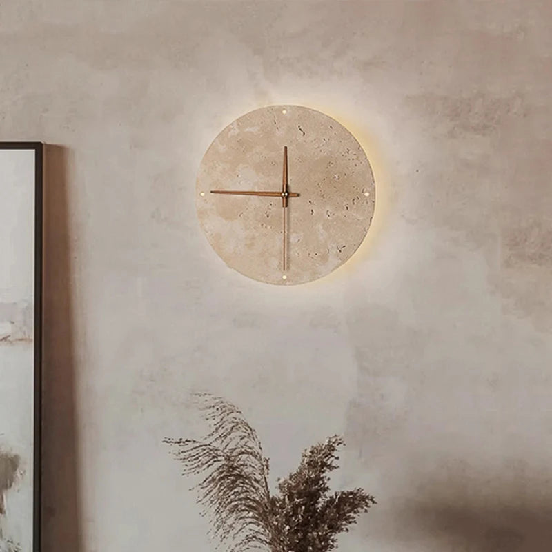 Modern wall clock for living room - Torwyn: A 9-inch travertine clock with brass hands and an LED halo, set against a neutral wall with dried grasses below.