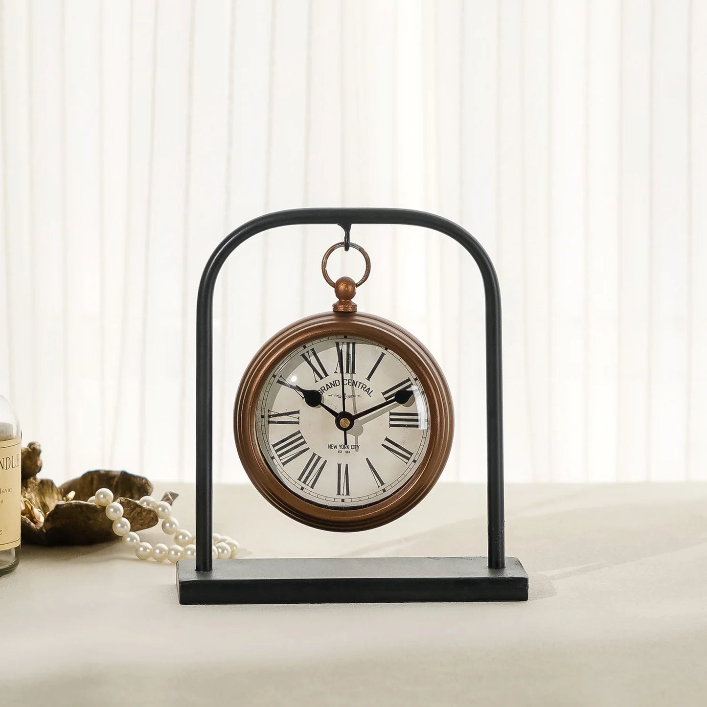 Retro Table Clock - Kelros: A vintage-style tabletop clock resembling a pocket watch, with Roman numerals, suspended by a black metal arch, alongside a gold-tone dish and glass jar. Perfect for desks.