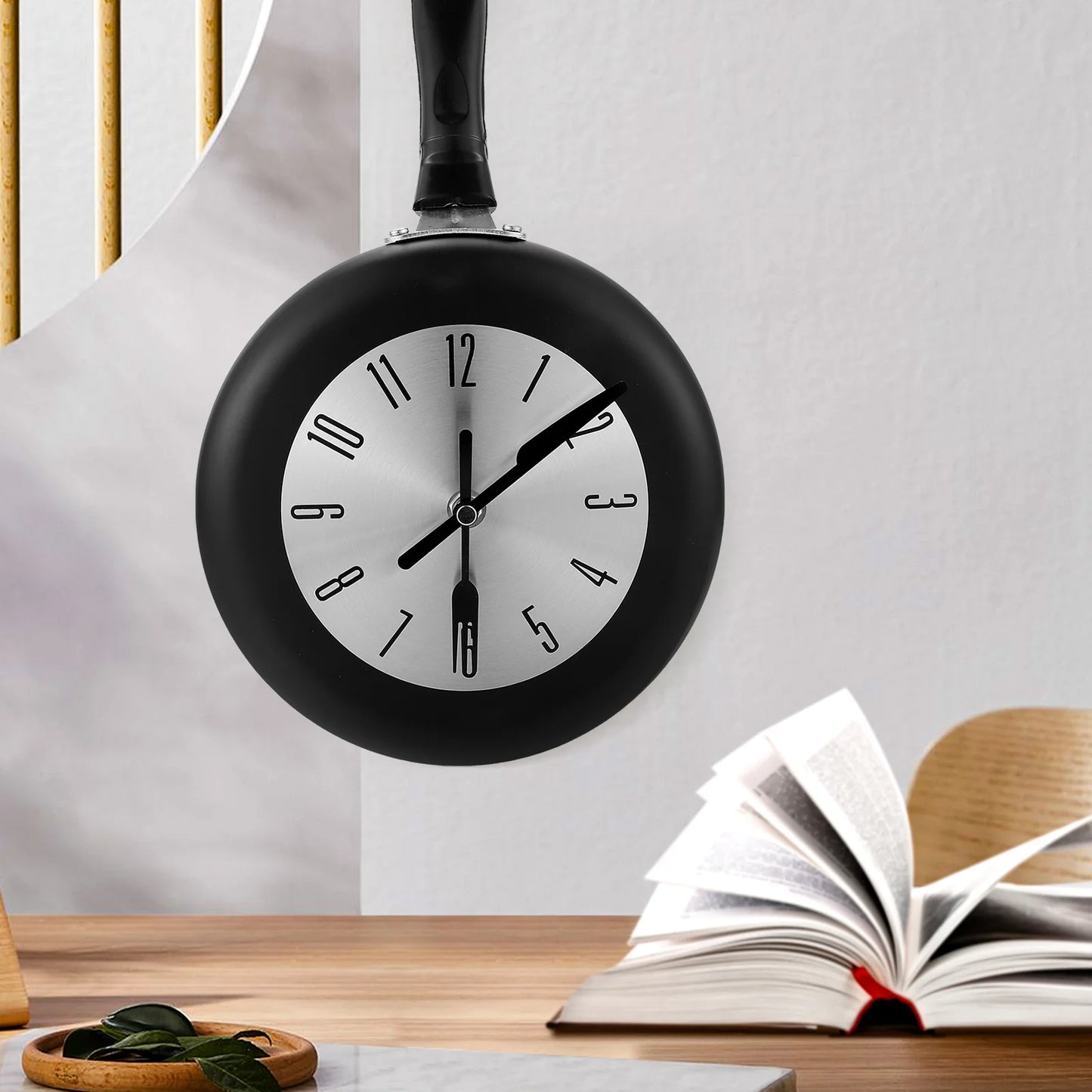 Frying Pan Wall Clock - Ashford: A novelty clock resembling a frying pan with a dial face and utensil-shaped hands, adding culinary charm. Mounted on a wall beside a table with an open book.