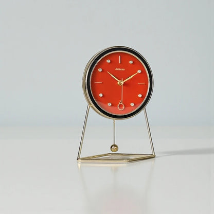 The Pendulum Table Clock - Dralen features a red dial, gold-tone hands, and a minimalist gold-metal stand with a swinging pendulum, blending classic and modern design elements.
