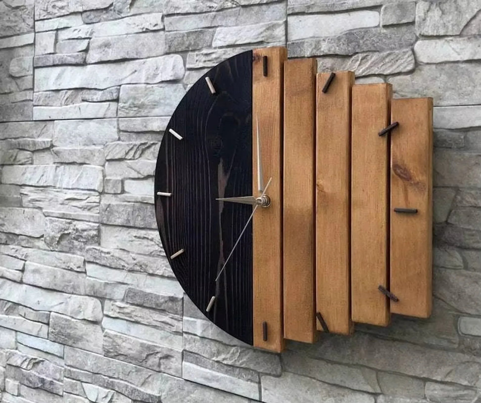 Modern wood wall clock - Clevor: This handcrafted clock features a black-stained semicircle and vertical wooden slats, accented with metal tick marks and silver hands, mounted on a textured stone-tile wall.