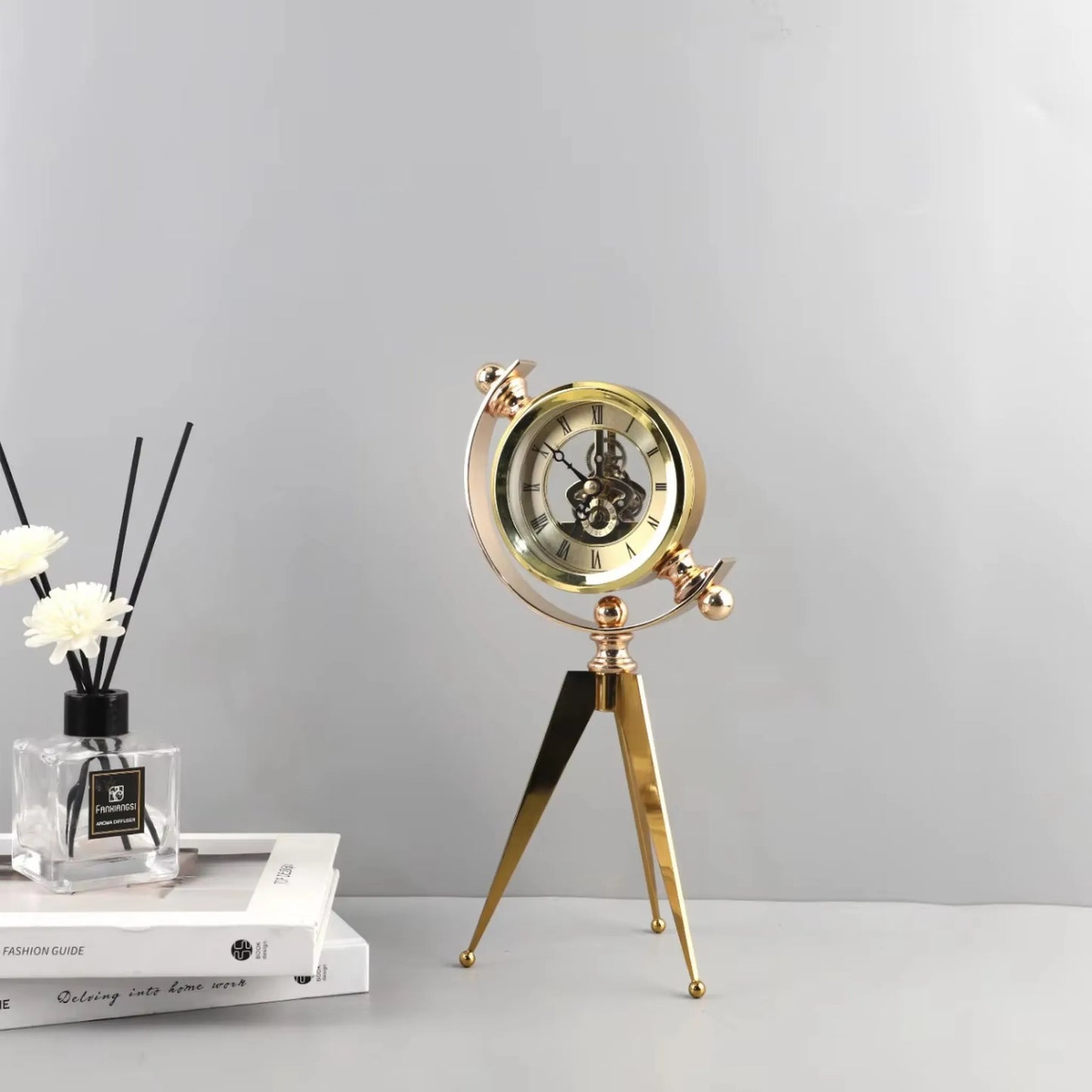 Vintage Table Clock - Gorven: A polished brass-colored, tripod-legged desk clock with exposed skeleton movement and Roman numerals, paired with a glass diffuser and books, offering vintage charm and functionality.