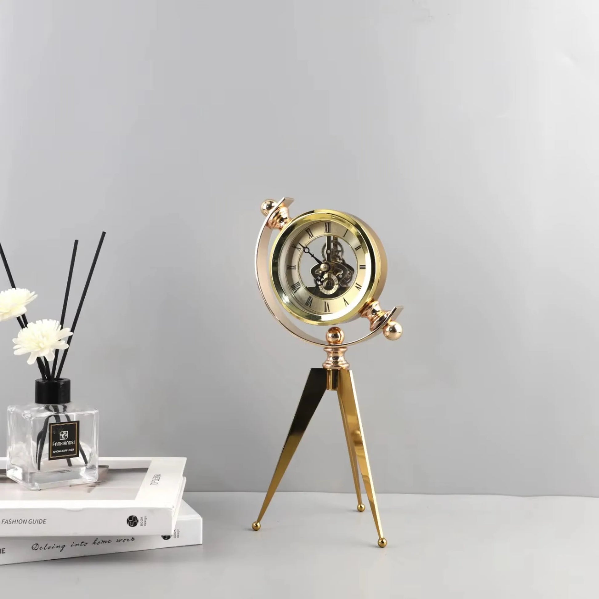 Vintage Table Clock - Gorven: A polished brass-colored, tripod-legged desk clock with exposed skeleton movement and Roman numerals, paired with a glass diffuser and books, offering vintage charm and functionality.