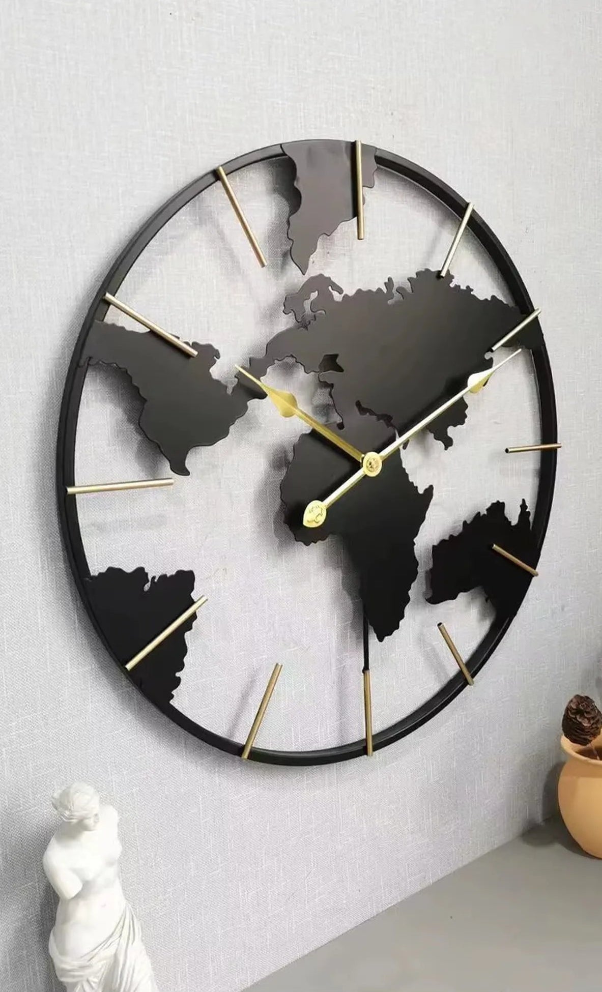 World Map Wall Clock - Claverton: A 23.6 black metal clock with continent silhouettes and gold-tone markers, elegantly mounted above a tabletop with a statuette, perfect for sophisticated living spaces.