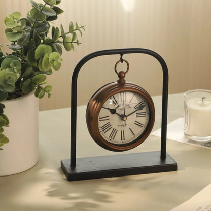 Retro Table Clock - Kelros, featuring a vintage design with Roman numerals and a brass-finished frame, elegantly suspended on a black metal arch, accompanied by a white potted plant and candle.
