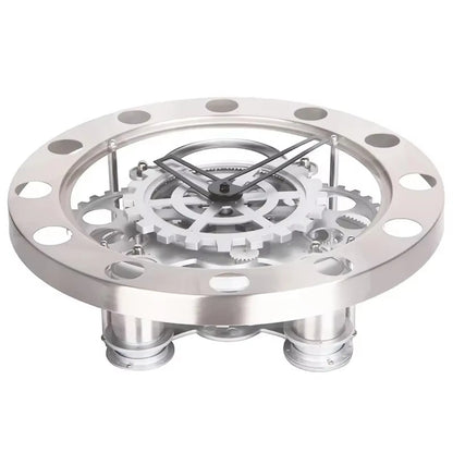 Modern Wall Clock Design - Elverton: A tabletop skeleton clock featuring a stainless-steel bezel with cut-outs, exposed interlocking gears, and black metal hands, elevated on short metal legs for a striking mechanical display.