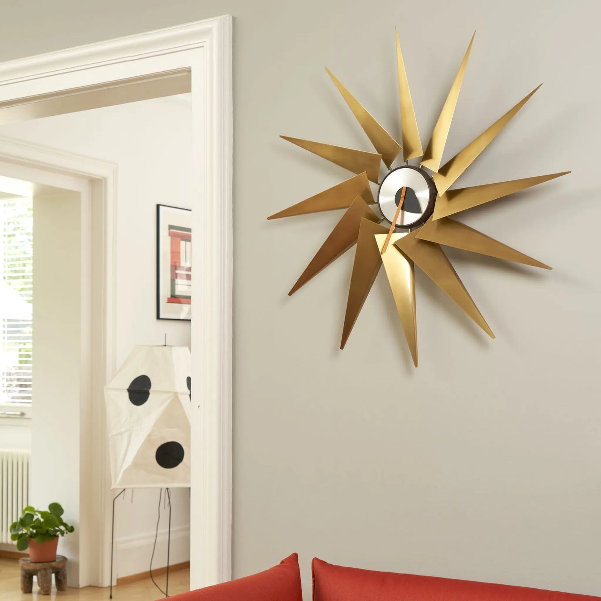 Orwick Golden wall clock on a wall in a room with a red couch and plant.