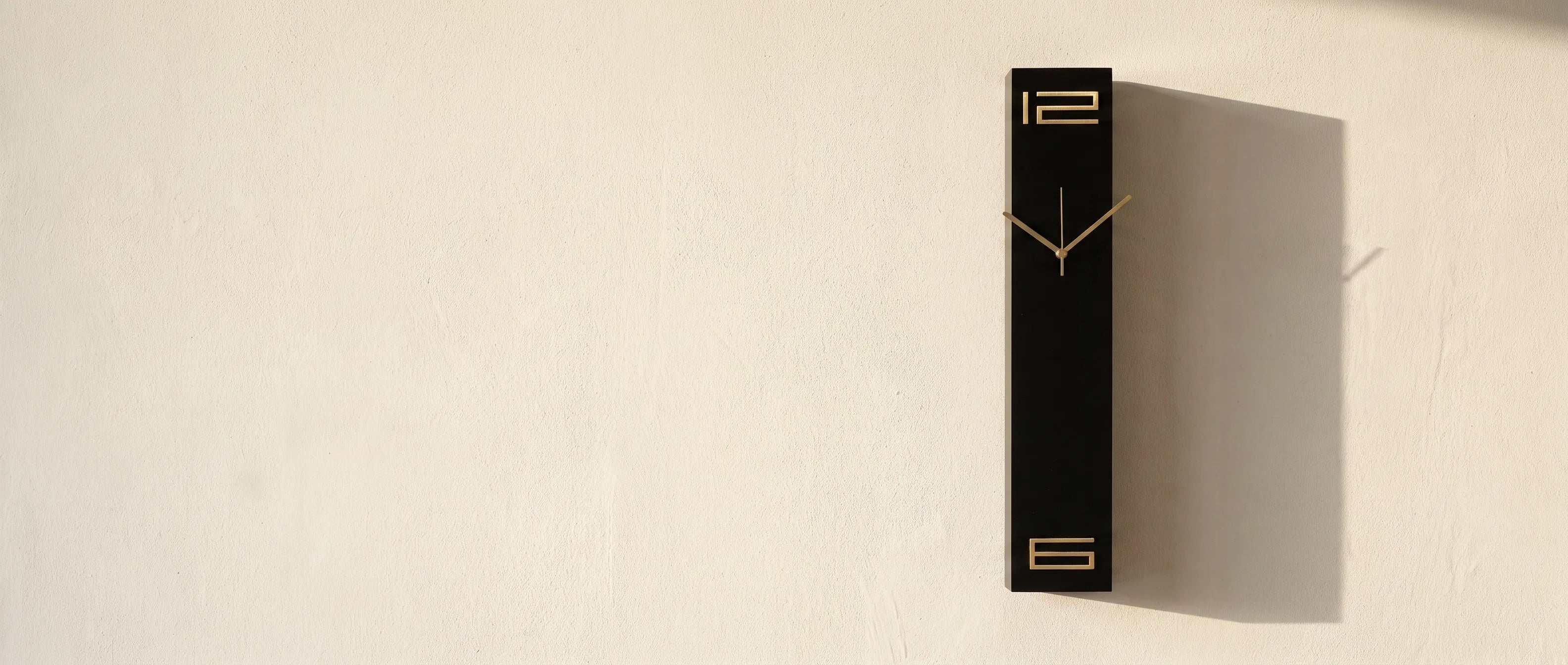 Black rectangular wall clock with brass numerals on a cream plaster wall in raking light